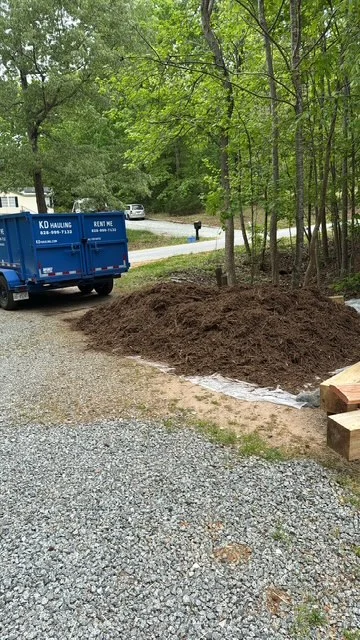 Bulk mulch delivery pile on gravel driveway with delivery truck and wooded residential property
