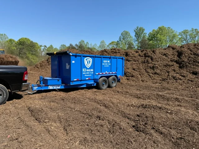 Blue dual-axle trailer loaded with dark mulch and soil materials at job site